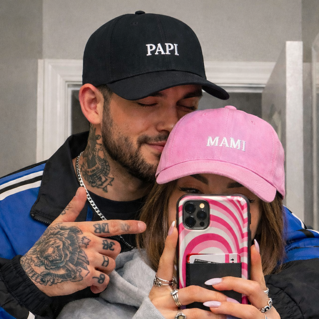 Two people taking a selfie, one wearing a black 'PAPI' cap and the other a pink 'MAMI' cap, with a phone displaying a pink and black pattern.
